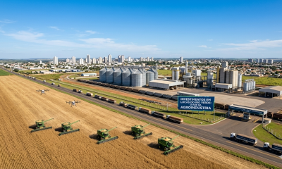 Vista aérea de colheitadeiras e drones operando em lavoura de Lucas do Rio Verde, Mato Grosso, com complexo industrial de grãos e silos ao fundo.