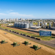Vista aérea de colheitadeiras e drones operando em lavoura de Lucas do Rio Verde, Mato Grosso, com complexo industrial de grãos e silos ao fundo.